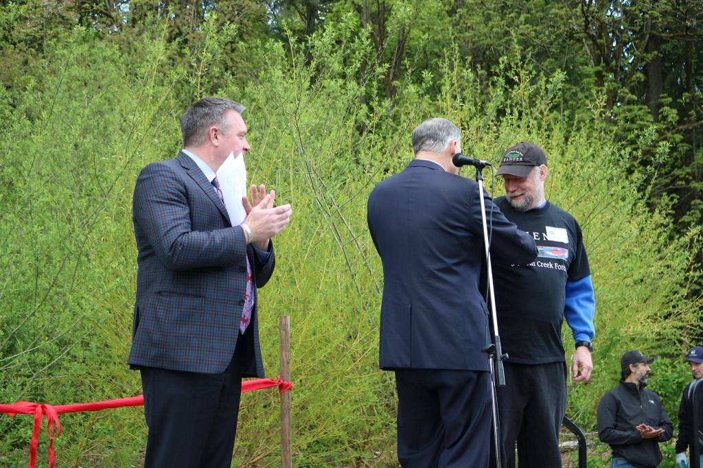 As part of being named Washingtonian of the Day, Gov. Jay Inslee (center) gives David Bain (right) the Washington State pin from his lapel. Bothell Mayor Andy Rheaume (left) applauds Bain receiving the honor on behalf of all of those who contributed to the city&rsquo;s acquisition of North Creek Forest. CATHERINE KRUMMEY / Bothell Reporter